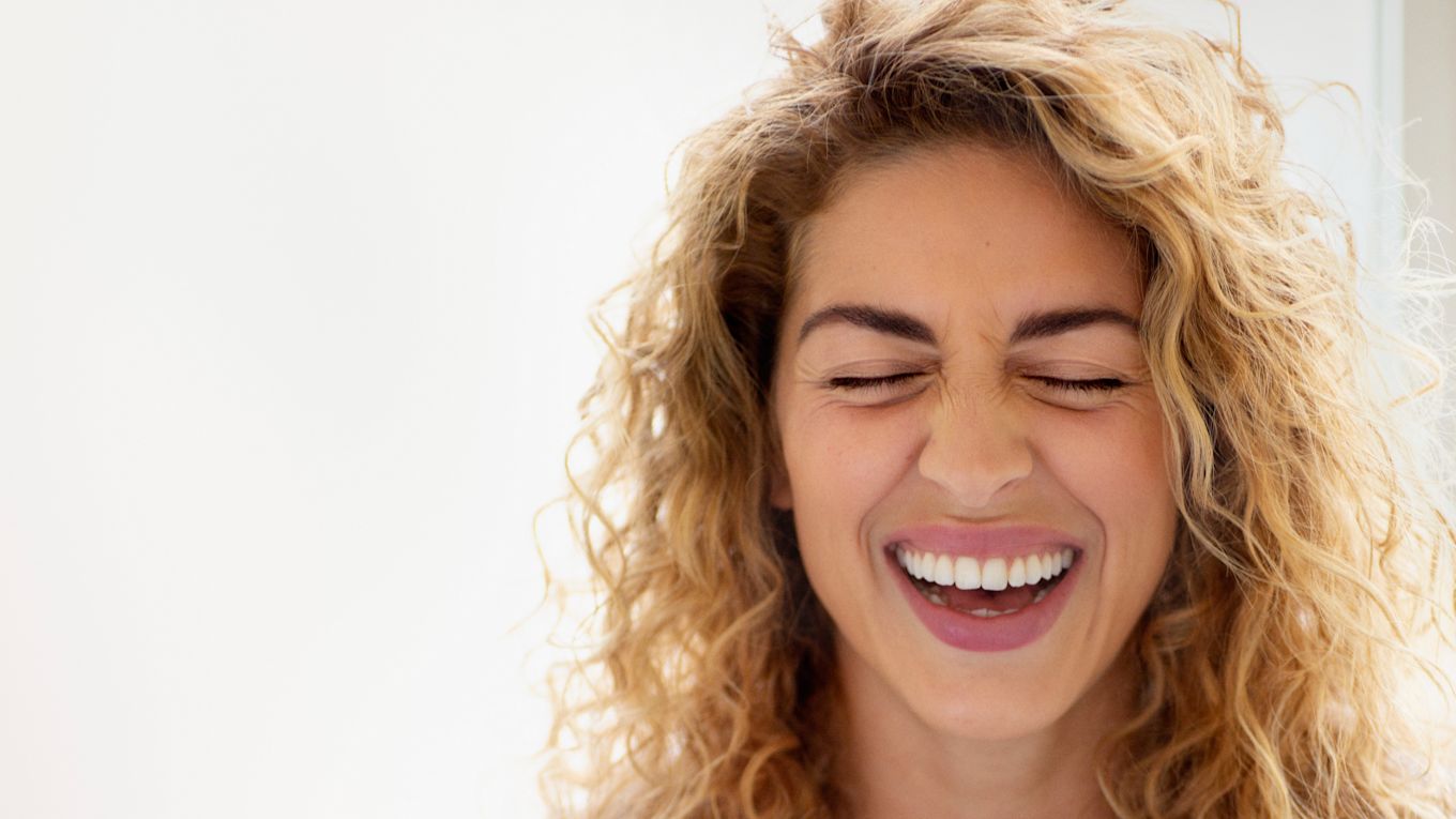 En kvinna med blont lockigt hår skrattar med slutna ögon.