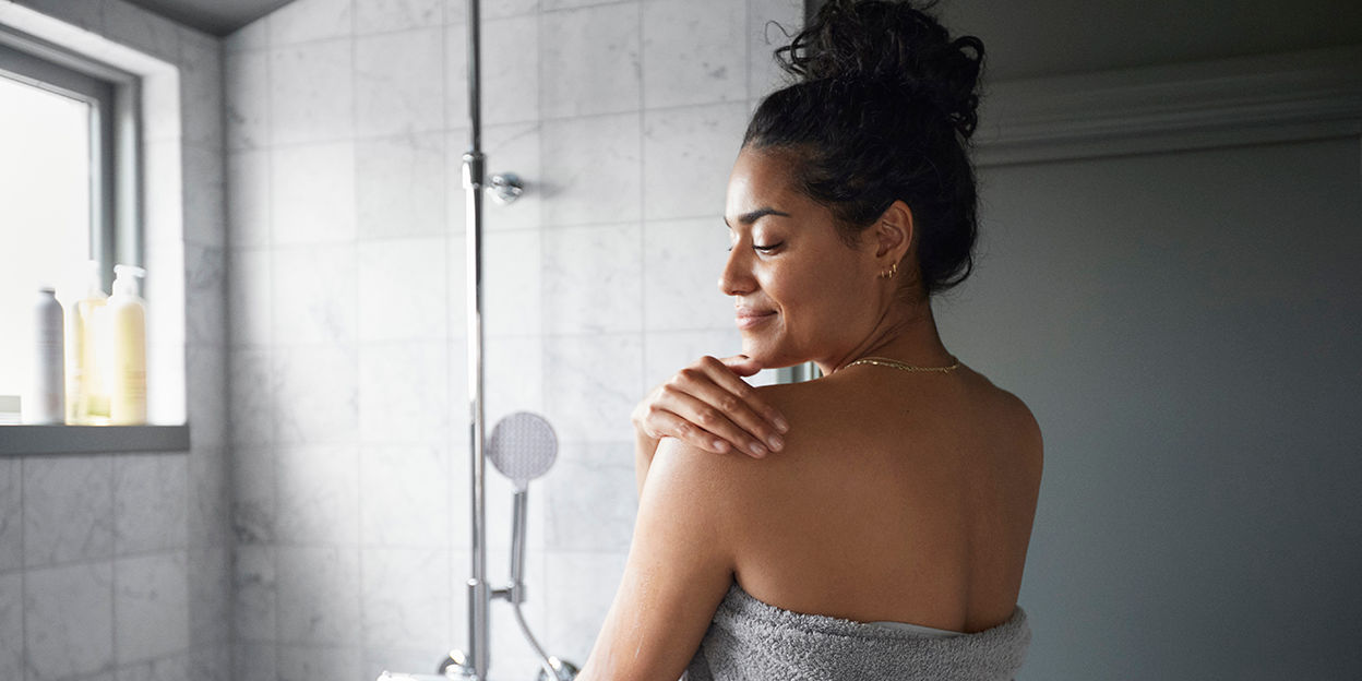 Kvinna står i duschen med handduk runt sig. 