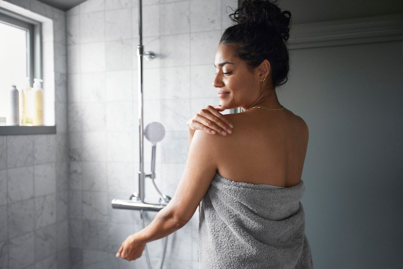 Kvinna står i badrum, invirad i handduk och smörjer in sin axel med lotion.