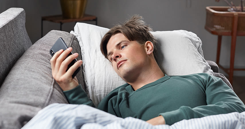 En man ligger i soffan och vilar med täcke och kudde. Han tittar på sin mobiltelefon som han håller med ena handen.