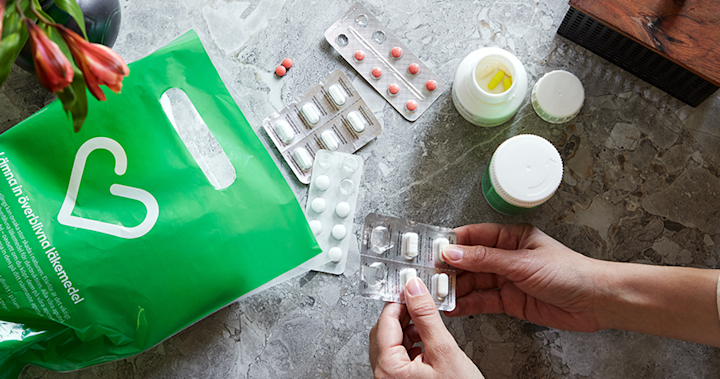 På ett grått marmorbord ligger röda, gula och vita tabletter och kapslar i burk och blister. Ett handpar håller i en tablettkarta med vita tabletter. En grön Apotek Hjärtat påse med texten: Lämna in överblivna läkemedel, är placerad bredvid är placerad bredvid. 