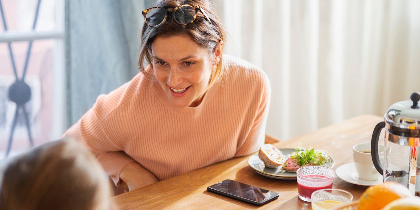 Mamma och barn sitter och äter frukost tillsammans och pratar. På bordet står tallrikar med frukost och smörgåsar, samt kaffe och smoothies.