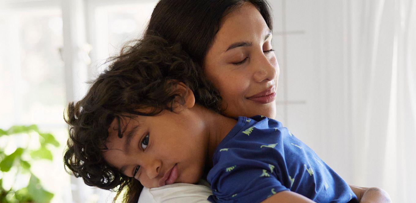 Mamma bär sin trötta son. Sonen vilar sitt huvud mot mammas axel.