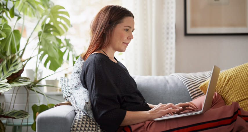 En kvinna sitter i soffan och knappar på datorn hon har i knät.
