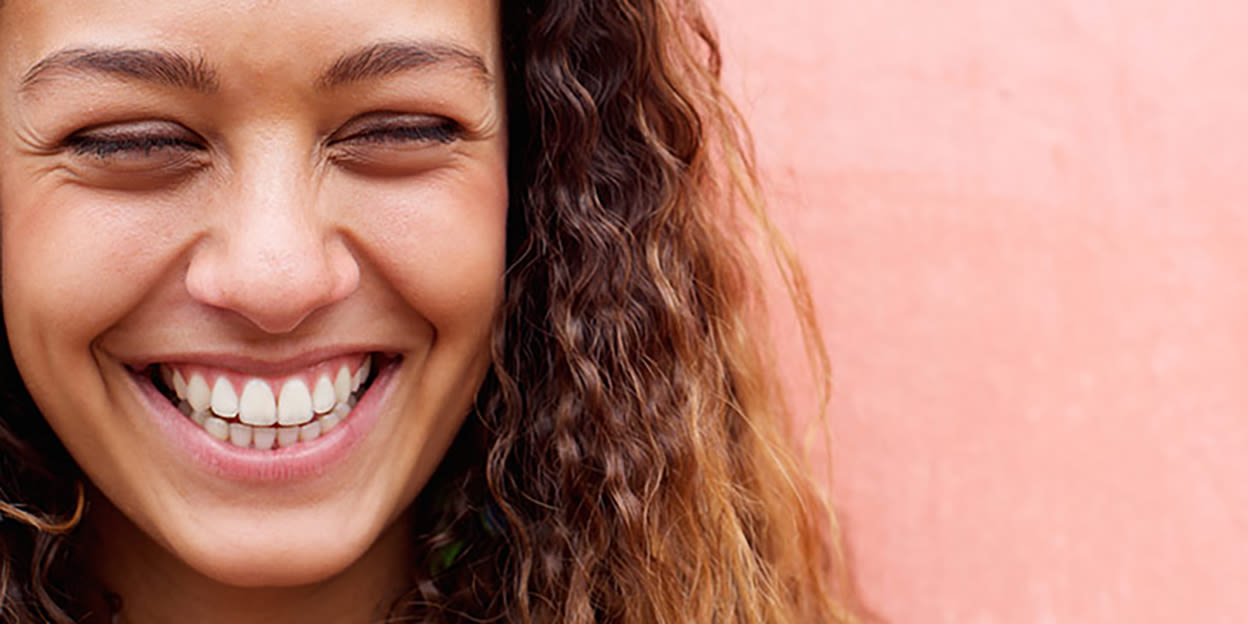 En långhårig tjej ler stort mot en rosa bakgrund.