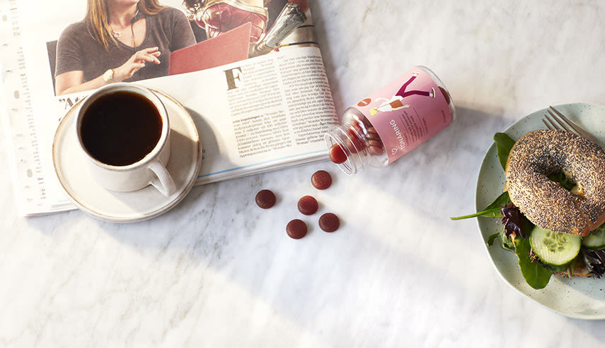 På ett vitt stenbord ligger en burk med Apotek Hjärtats multivitamintabletter för tonåringar. Bredvid på bordet ligger en tidning, en kaffekopp och en tallrik med en bagel på.