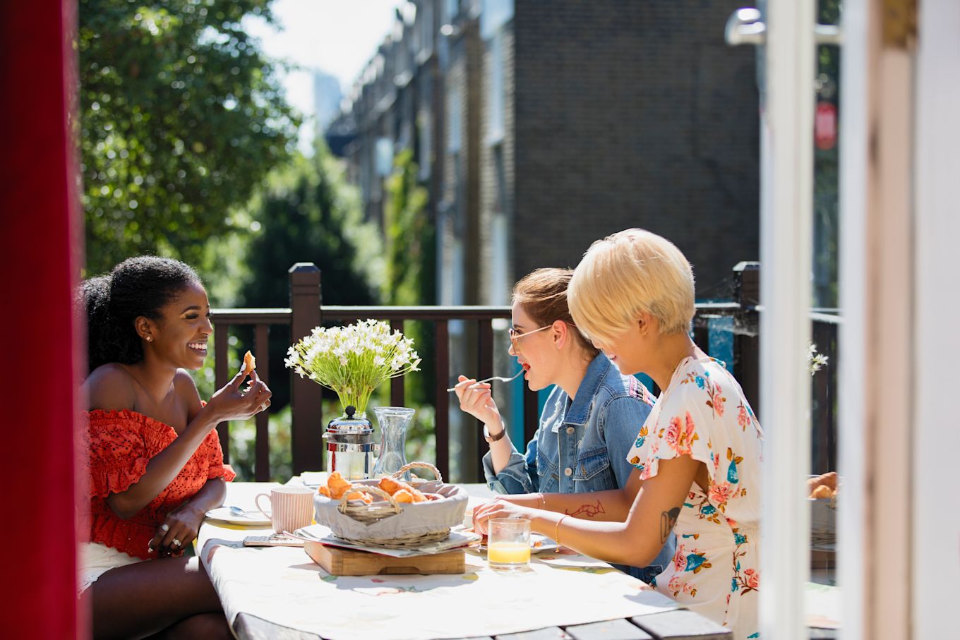 Tre unga kvinnor sitter på en balkong i solen och äter mat tillsammans och skrattar.
