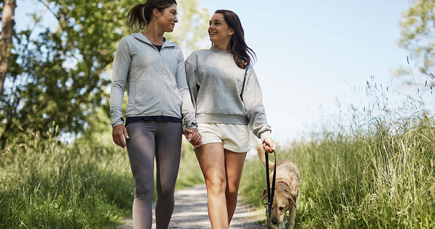 Två kvinnor går hand i hand på en gräsbevuxen stig och ler mot varandra, omgivna av träd och en klarblå himmel.
