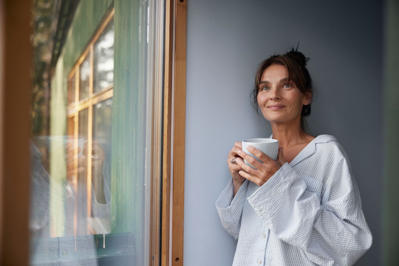 Kvinna står vid ett fönster, iklädd pyjamas med en temugg i händerna. Hon tittar åt sidan och ser nöjd ut.