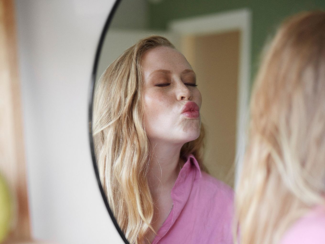 Sminkad kvinna i rosa tröja står och speglar sig och plutar med läpparna.