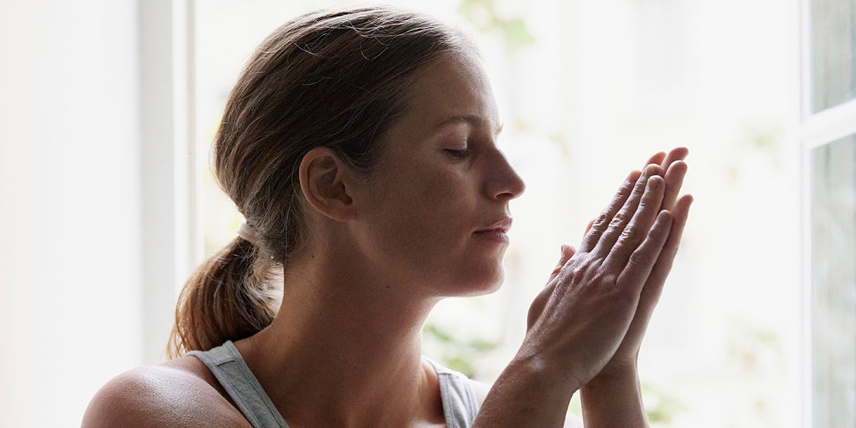 Kvinna i profil som kollar snett nedåt och håller ihop sina händer. Hon står framför ett fönster. 