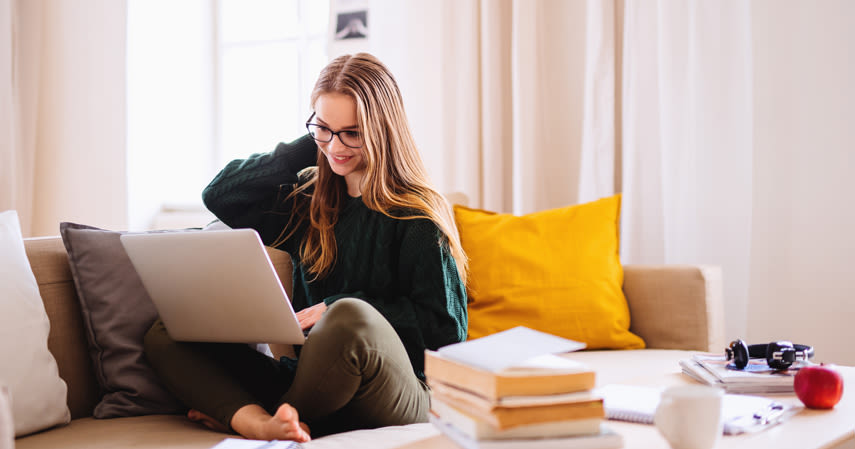 En kvinna sitter i soffan och jobbar på en laptop. Bredvid henne på soffbordet ligger en hög med böcker, en kaffekopp, hörlurar och ett äpple.