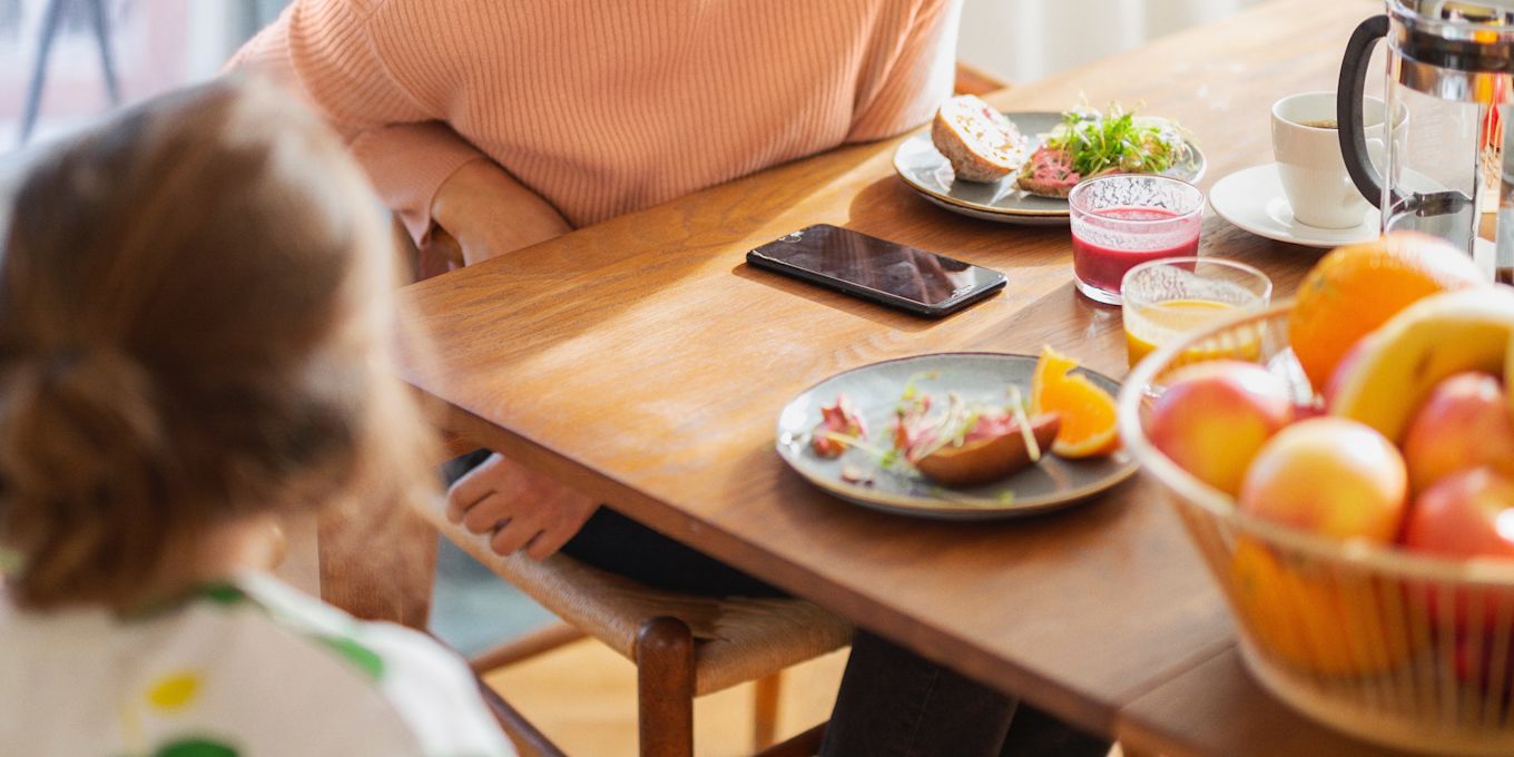 Ett frukostbord fyllt med olika frukt, grönsaker, mackor, kaffe och smoothies. Mamma och ett barn sitter vid bordet och äter och pratar.