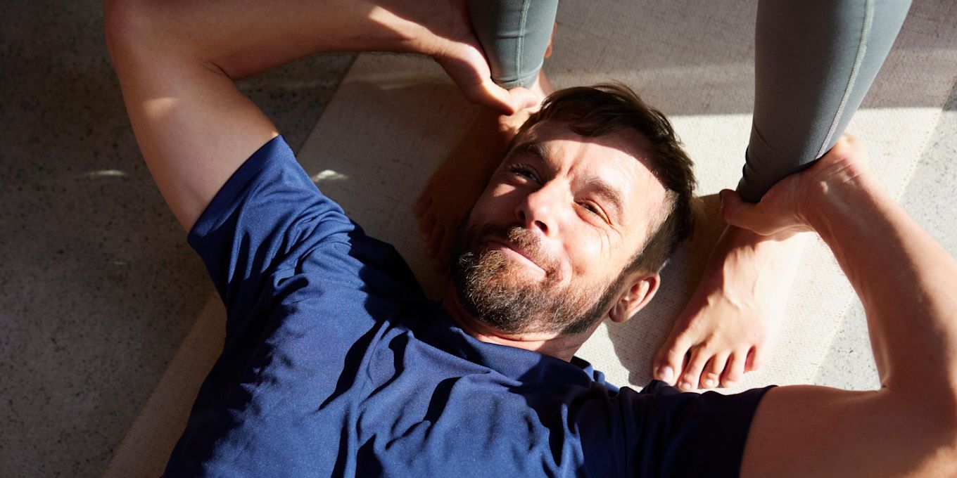 Man ligger på en yogamatta på golvet och tränar, vid hans huvud står en kvinnas fötter.