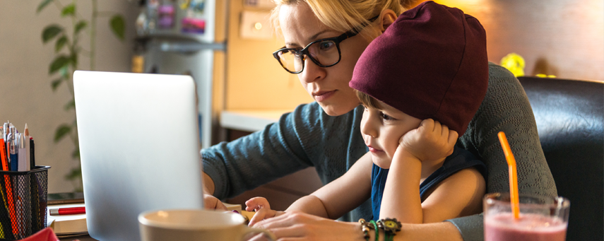Förälder har sitt barn i knät och de båda tittar på en laptop. På bordet finns en smoothie, kaffekopp och pennor.