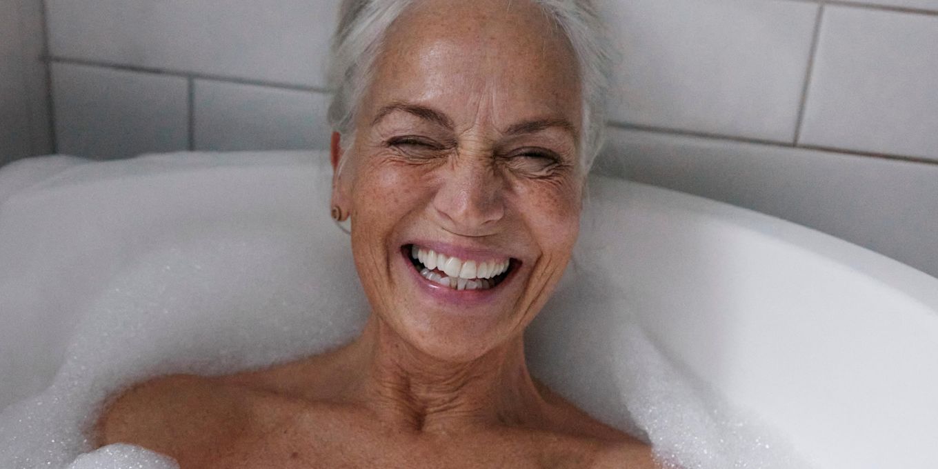 Äldre kvinna ligger i badkaret med bubblor runtomkring sig och skrattar.