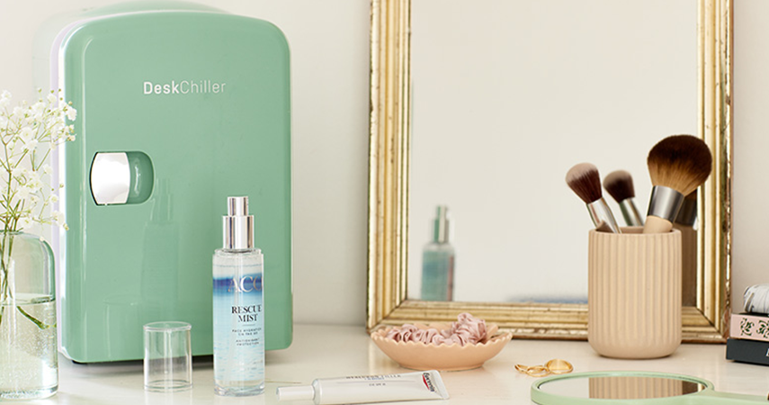 På ett sminkbord står en liten grön kyl för hudvård, olika skönhetsprodukter, sminkborstar och en spegel.