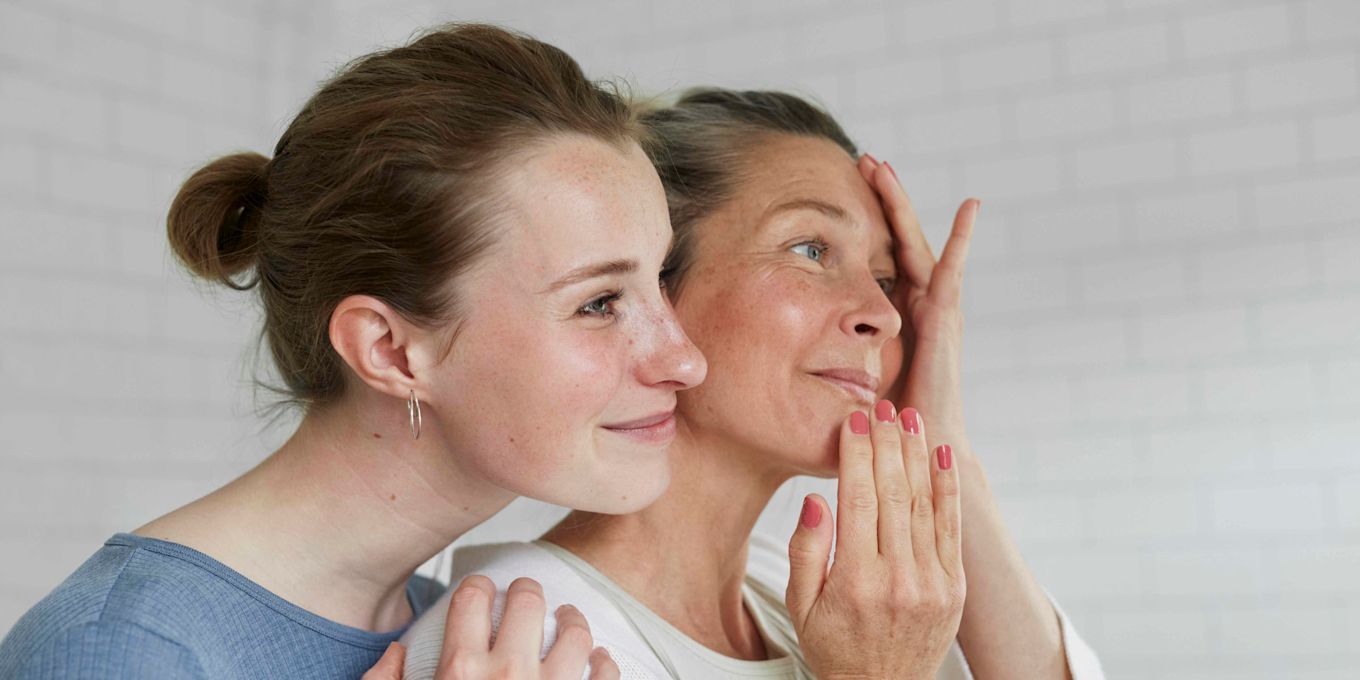 Två kvinnor kollar bort från kameran och ler. Ena kvinnan står lite bakom och håller om den andra kvinnan. 