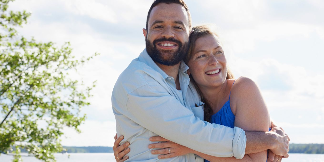 Glad man och kvinna står vid vattnet i sommarsolen och håller om varandra.