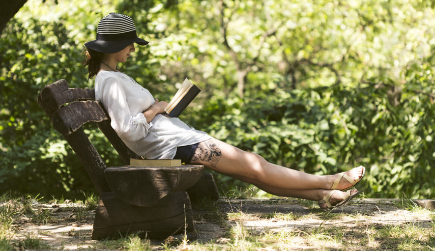 En kvinna i hatt sitter på en träbänk och läser en bok i en grönskande park. Solljuset filtrerar genom träden.