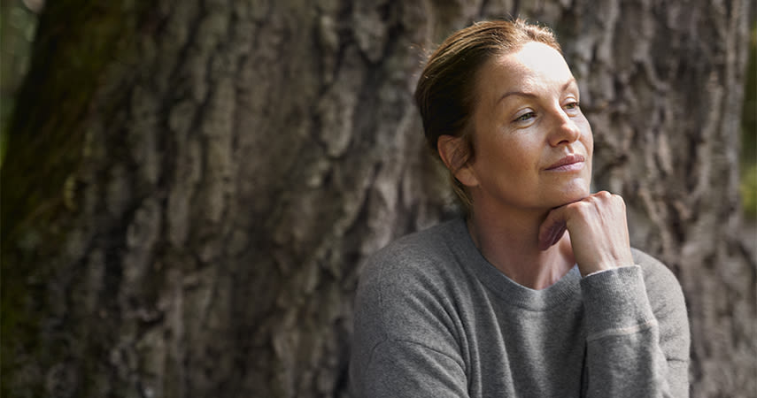 Kvinna sitter utomhus framför ett stort träd och tittar åt sidan.