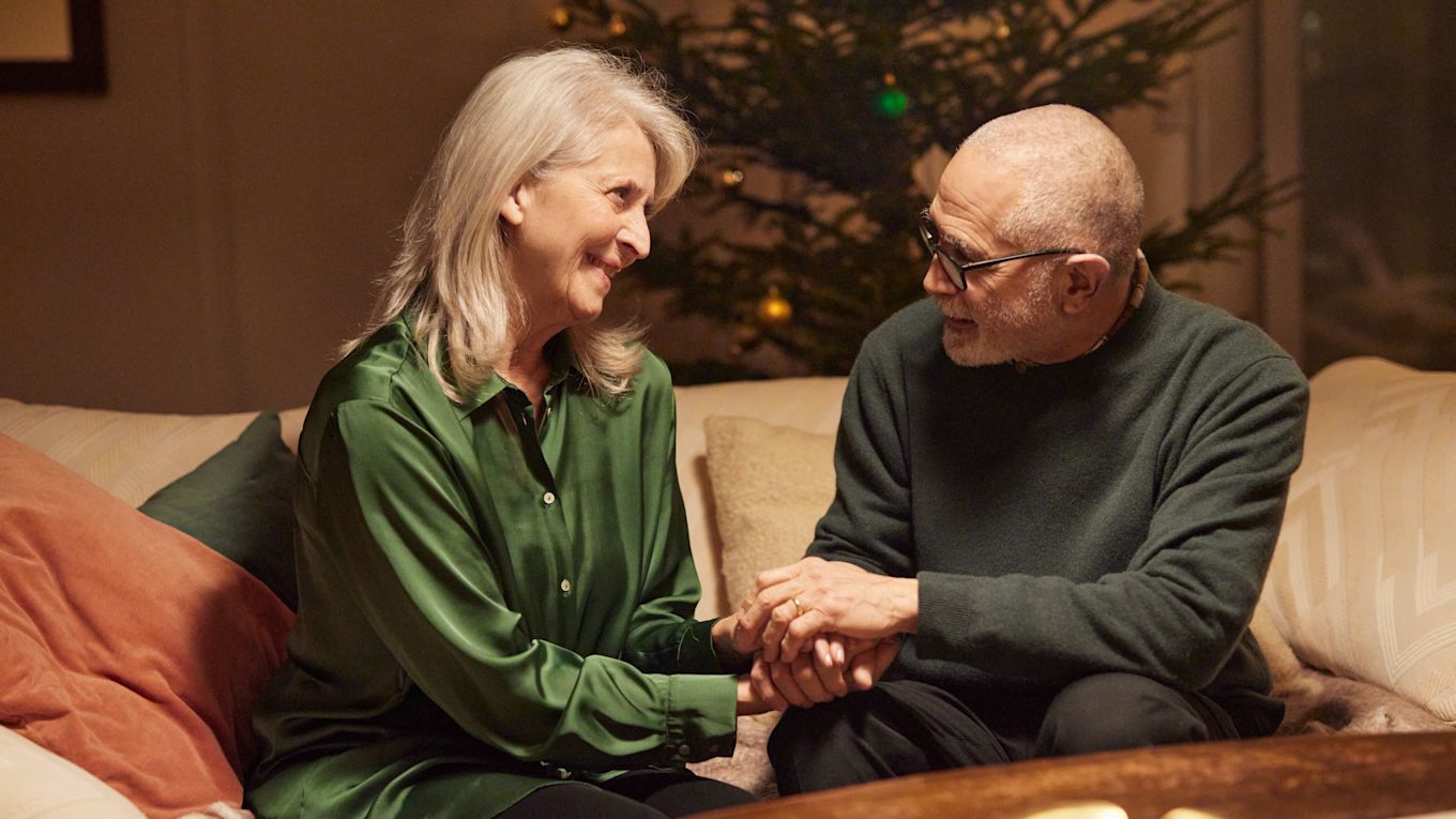 En äldre man och kvinna sitter tillsammans i en soffa med en julgran i bakgrunden. De håller varandra i händerna och ler varmt mot varandra.
