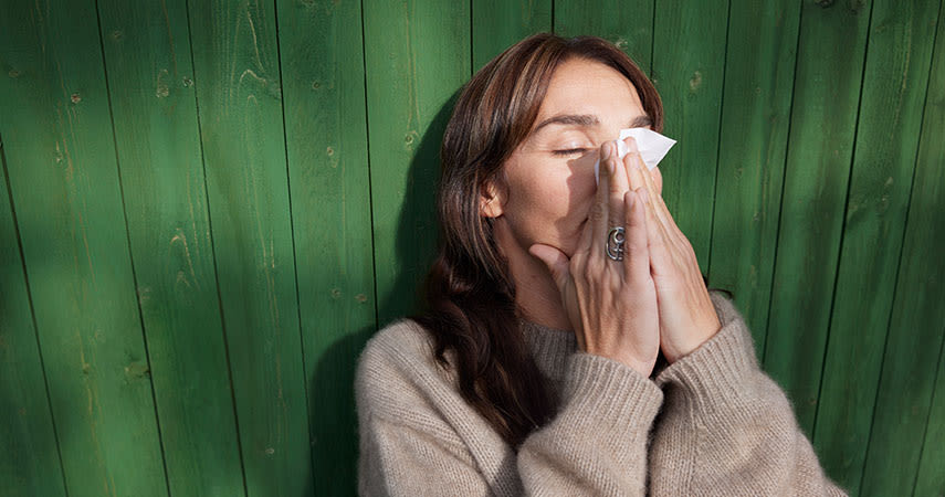 Kvinna sitter utomhus, lutad mot en grön vägg och snyter sig.