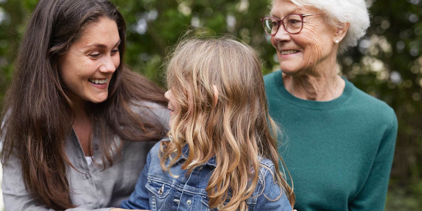 Mormor, mamma och dotter sitter utomhus tillsammans och kramas och skrattar.