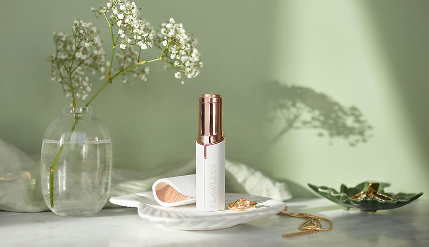 En Flawless Deluxe Facial Hair Remover står på ett snäckformat fat i ett badrum. Bredvid står en liten vas med vita blommor och en guldfärgad kedja ligger på andra sidan av fatet.